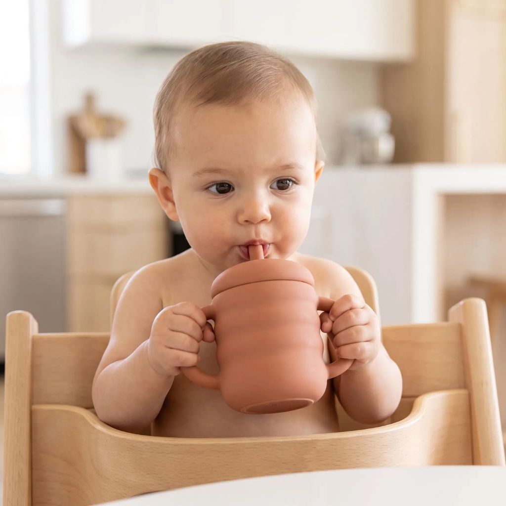 Bébé tenant sa tasse d'apprentissage par les deux anses et buvant à la paille